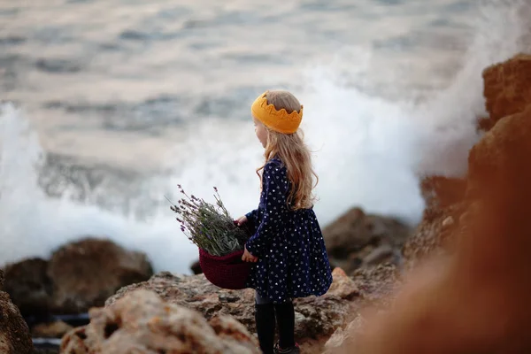 Una chica guapa con una corona de punto amarillo sostiene una canasta de cordón en sus manos y se para en la orilla del mar —  Fotos de Stock