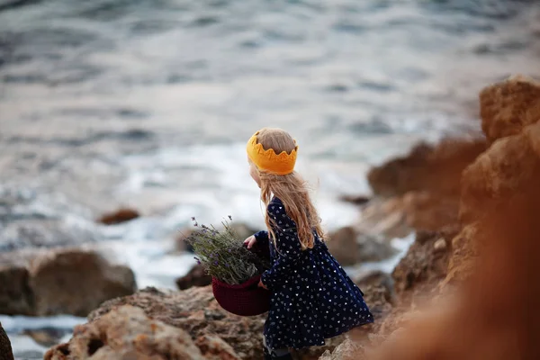 Uma menina bonita em uma coroa de malha amarela segura uma cesta de corda em suas mãos e fica na beira-mar — Fotografia de Stock