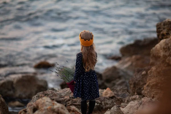 Una chica guapa con una corona de punto amarillo sostiene una canasta de cordón en sus manos y se para en la orilla del mar —  Fotos de Stock