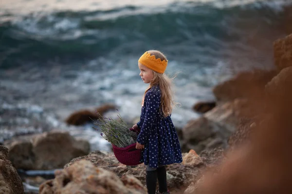 Ein hübsches Mädchen in einer gelben Strickkrone hält einen Korb mit Kordel in der Hand und steht am Ufer des Meeres — Stockfoto