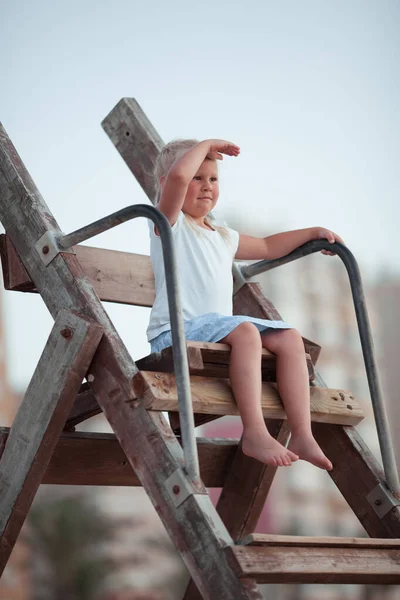 Pretty little girl sits on a lifeguard chair and looks into the distance — Stock Photo, Image
