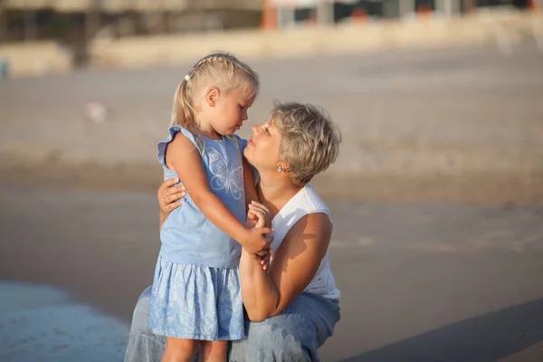 Portret van een gelukkige moeder en een kleine dochter. Ze knuffelen op de achtergrond van de zee. — Stockfoto