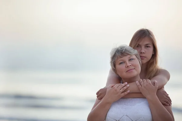 Portret van een gelukkige moeder en volwassen dochter. Ze knuffelen op de achtergrond van de zee. — Stockfoto