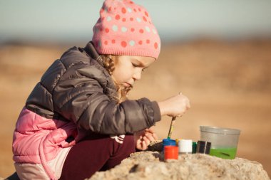Kız deniz kenarında çakıl taşları çiziyor, açık havada.