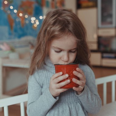 Güzel küçük kız sıcak bir odada, yatağında kırmızı bir kupayla kakao içiyor.