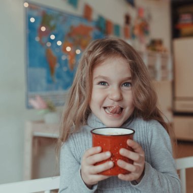 Güzel küçük kız sıcak bir odada, yatağında kırmızı bir kupayla kakao içiyor.