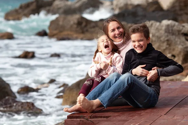快乐的妈妈 大儿子和小女儿赤脚坐在木制的码头上 在大海和岩石的背景下 在户外嬉笑 母亲与儿童之间的交流 — 图库照片