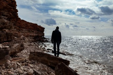 Yetişkin bir adam denize bakar, sırtı dönük durur ve ceket ve kot pantolon giyer, baharda deniz kıyısında yürür, siluet, açık havada