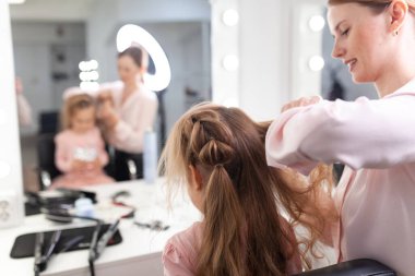 Parlak bir kuaförde saç stili üzerine çalışan kız, küçük, güzel bir modelden bukleler ve örgüler yapıyor.