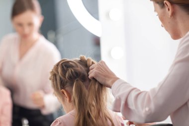 Parlak bir kuaförde saç stili üzerine çalışan kız, küçük, güzel bir modelden bukleler ve örgüler yapıyor.