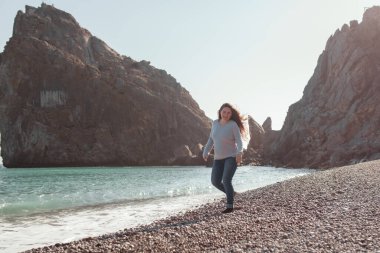 Mutlu güzel kız deniz kıyısı boyunca yürür. Deniz ve kayaların arkasında. Güzel bir manzaranın arkasından.