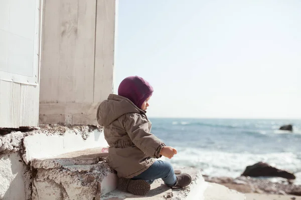 一个女孩坐在一座由白色木板制成的建筑旁边的石阶上 婴儿身穿帽子和夹克 望着冬季的大海 — 图库照片