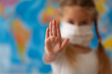 Yüzünde maske olan küçük bir kız. Koronavirüs kavramı ve bakterilere ve kirli havaya karşı koruma. Çocuğun eli dünya haritasının arka planında bir durak gösterir