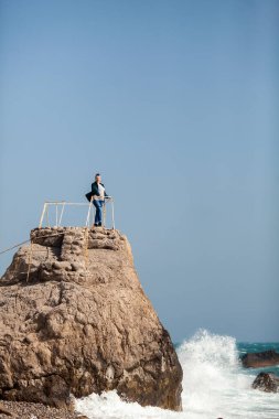Bir kadın büyük bir kayanın kenarında duruyor ve fırtınalı denize bakıyor.