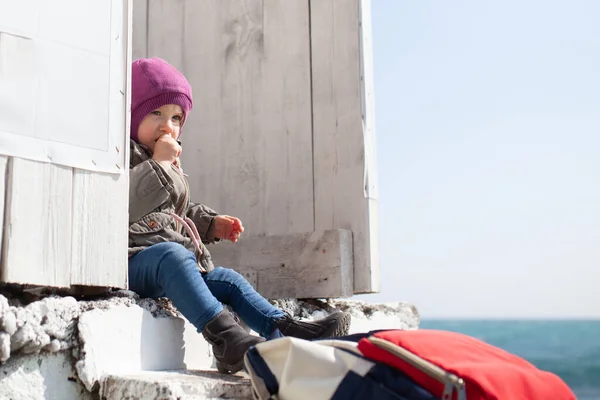 一个女孩坐在一座由白色木板制成的建筑旁边的石阶上 婴儿身穿帽子和夹克 望着冬季的大海 — 图库照片