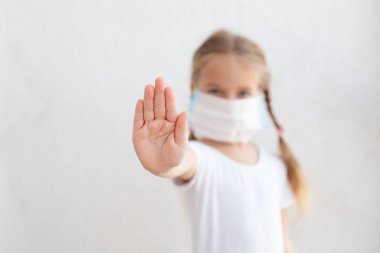Yüzünde maske olan küçük bir kız. Koronavirüs kavramı ve bakterilere ve kirli havaya karşı koruma. Çocuk eli beyaz bir duvar arkaplanında durur