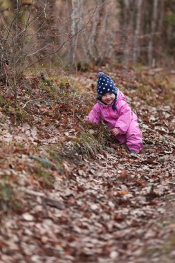Küçük bir çocuk sonbahar ormanında yürür, pembe tulum ve şapka giyen bir kız, temiz orman havası çocukların sağlığı için iyidir, mutlu bebek kurumuş yaprakların üzerine oturur.