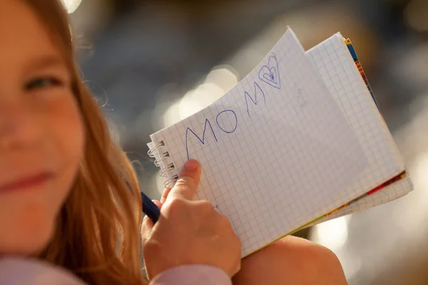 Παιδικό Χέρι Γράφει Ένα Σημειωματάριο Λέξη Μαμά Ένα Κορίτσι Κάθεται — Φωτογραφία Αρχείου