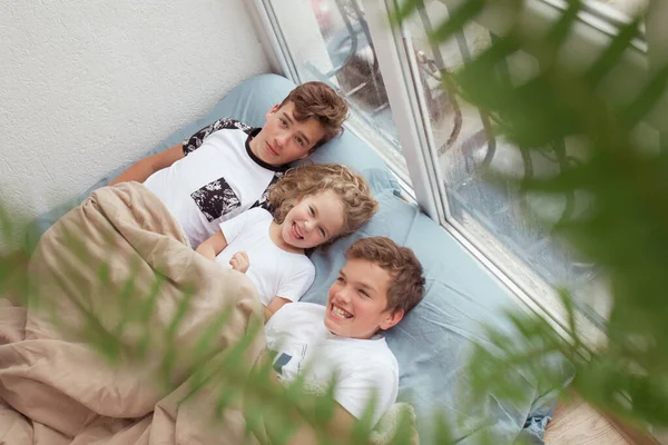 小女孩和哥哥们躺在窗边的沙发上 孩子们在屋里笑着和说话 — 图库照片