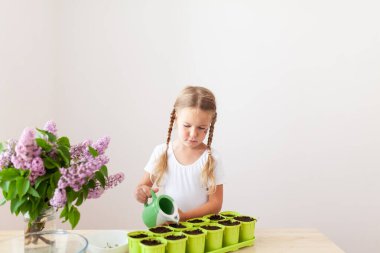 Beyaz tişörtlü küçük bir kız yeşil saksılara bezelye tohumu ekiyor, bir çocuk bitkilerle ilgileniyor, pencerenin kenarında bir ev bahçesi var.