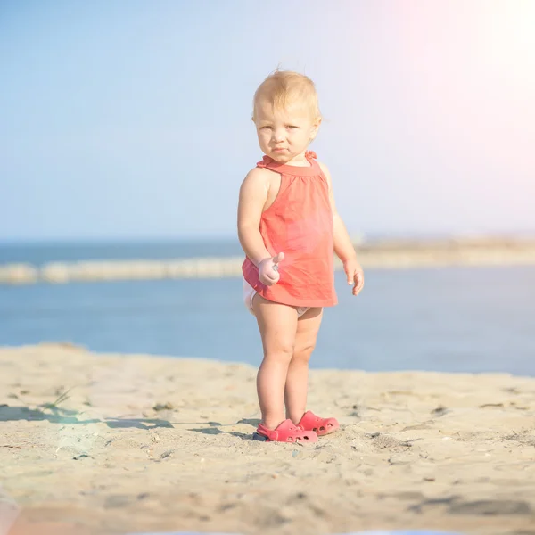 Baby girl in red dress playing on sandy beach near the sea. — Stock Photo, Image