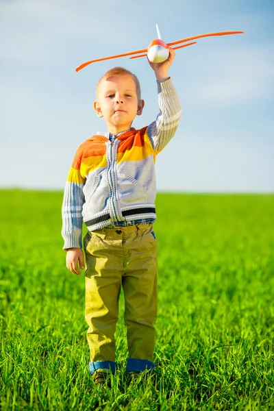 Gelukkige jongen spelen met speelgoed vliegtuig tegen blauwe zomer hemel en groen veld achtergrond. — Stockfoto