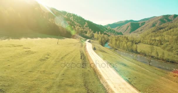 Luchtfoto van de Uhd 4k. Lage air-vlucht over de bergweg van de landelijke vuil en weiland op de zonnige Zomerochtend. In de buurt van de groene bomen, zonnestralen en berg rivier — Stockvideo