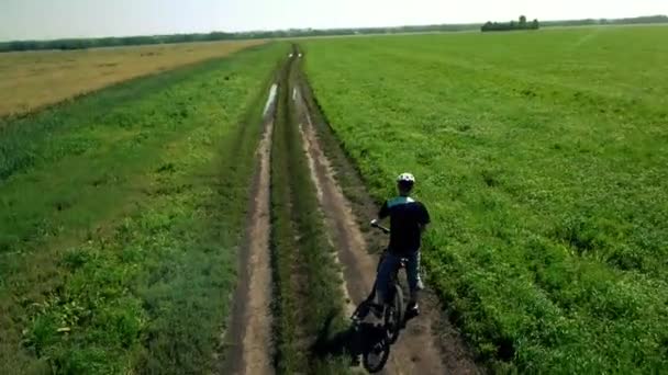 AERIAL: Jovem pedalando de bicicleta na estrada rural através do campo verde e amarelo . — Vídeo de Stock