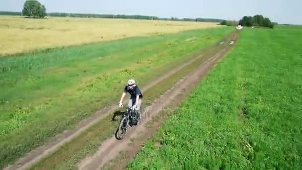 Antena: Młody człowiek na rowerze na rowerze na wiejskiej drodze poprzez zielone i żółte pola. — Wideo stockowe