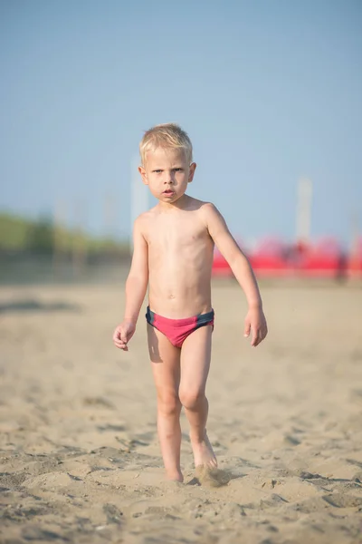 Ładny chłopczyk chodzenie na piaszczystej plaży w pobliżu morza. Na wybrzeżu Oceanu. — Zdjęcie stockowe