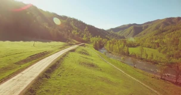 Vôo a meio do ar sobre o rio e o prado frescos da montanha na manhã ensolarada do verão. Estrada de terra rural abaixo. — Vídeo de Stock