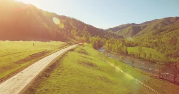 Mittlerer Luftflug über frischem Gebirgsfluss und Wiese am sonnigen Sommermorgen. Schotterpiste unten. — Stockvideo