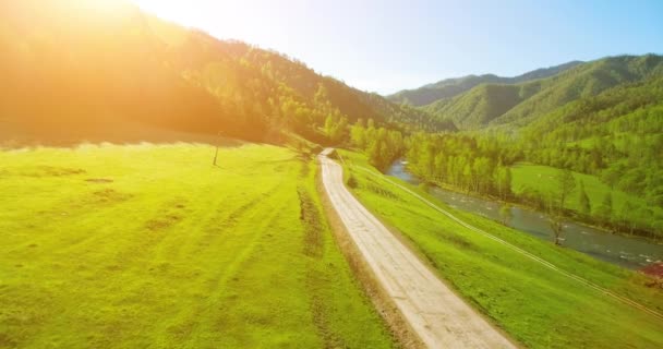 4 k Uhd 空撮。山農村未舗装の道路と晴れた夏の朝に草原上低空気飛行。近くには緑の木々、太陽光線、山川 — ストック動画