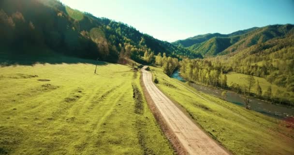 Widok z lotu ptaka Uhd 4k. Niski Lot nad górskich obszarów wiejskich polnej drodze i łąka w słoneczny letni poranek. W pobliżu zielonych drzew, promienie słońca i górskiej rzeki — Wideo stockowe