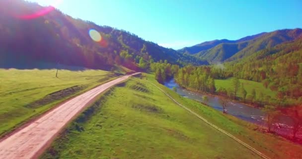 Mid air flight over fresh mountain river and meadow at sunny summer morning. Rural dirt road below. — Stock Video