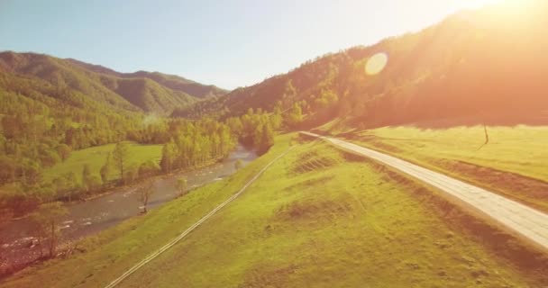 Mid air flight over fresh mountain river and meadow at sunny summer morning. Rural dirt road below. — Stock Video