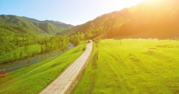 Aerial UHD 4K view. Low air flight over mountain rural dirt road and meadow at sunny summer morning. Near green trees, sun rays and mountain river — Stock Video