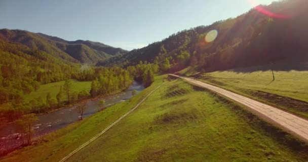 Vôo a meio do ar sobre o rio e o prado frescos da montanha na manhã ensolarada do verão. Estrada de terra rural abaixo. — Vídeo de Stock