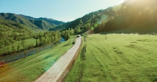 Vista aérea UHD 4K. Vôo de ar baixo sobre montanha estrada de terra rural e prado na manhã de verão ensolarado. Perto de árvores verdes, raios de sol e rio de montanha — Vídeo de Stock