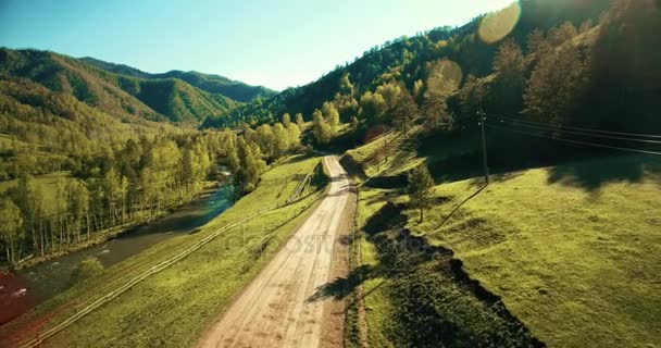 Vista aérea UHD 4K. Vuelo de aire bajo sobre carretera de tierra rural de montaña y prado en la soleada mañana de verano. Cerca de árboles verdes, rayos de sol y río de montaña — Vídeos de Stock