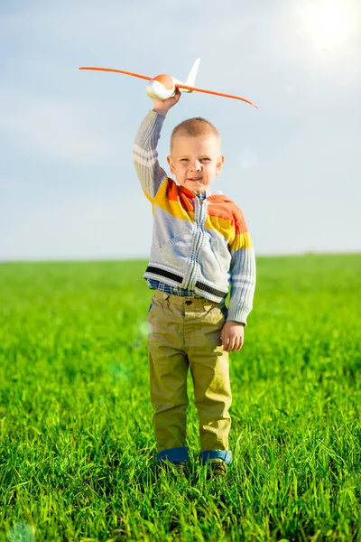 Gelukkige jongen spelen met speelgoed vliegtuig tegen blauwe zomer hemel en groen veld achtergrond. — Stockfoto