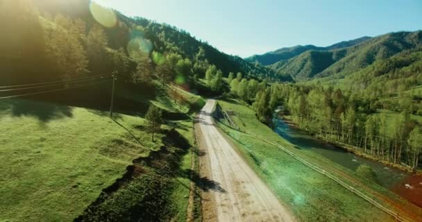 Uhd 4k légifelvételek. Alacsony légijárat hegyi vidéki földút és rét a napsütéses nyári reggel. Zöld fák, a nap sugarai és a hegyi folyó közelében — Stock videók