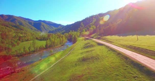 Vôo a meio do ar sobre o rio e o prado frescos da montanha na manhã ensolarada do verão. Estrada de terra rural abaixo. — Vídeo de Stock