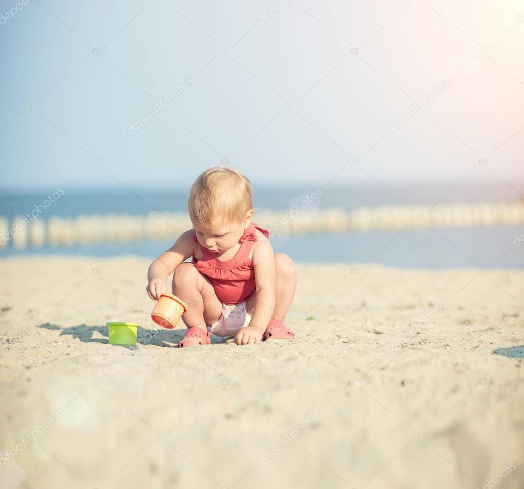 Baby girl in red dress playing on sandy beach near the sea. 