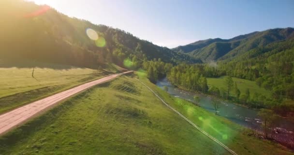 Vôo a meio do ar sobre o rio e o prado frescos da montanha na manhã ensolarada do verão. Estrada de terra rural abaixo. — Vídeo de Stock