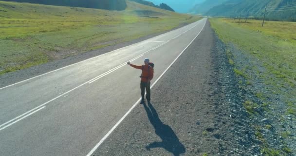 アスファルトの道路を歩くヒッチハイカーの観光客の上のフライト。夏の日に巨大な田舎の谷。リュックハイキング男. — ストック動画