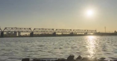 Şehir nehir kıyısının zaman çizelgesi. Güneş ışınları, mavi gökyüzü ve ufukta demiryolu köprüsü. Düz küçük dalgalar, su ve motorlu kaydırak hareketleriyle yaz günbatımı zamanı