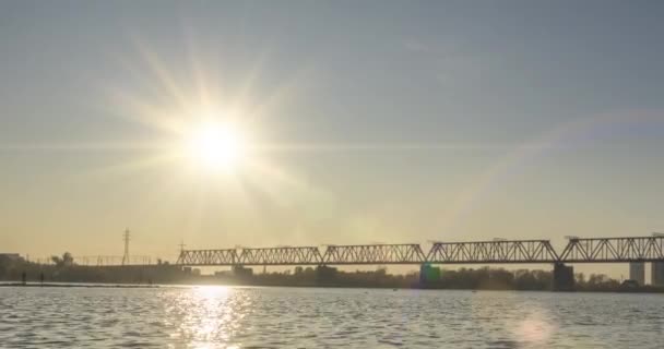 Timelapse de la orilla del río ciudad. Rayos solares, cielo azul y puente ferroviario sobre horizontal. Hora del atardecer de verano con olas planas y movimiento deslizante motorizado — Vídeos de Stock