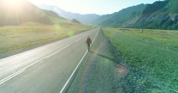 Vuelo sobre autoestopista turista caminando por carretera asfaltada. Gran valle rural en el día de verano. Mochila senderismo chico. — Vídeo de stock