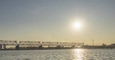 Şehir nehir kıyısının zaman çizelgesi. Güneş ışınları, mavi gökyüzü ve ufukta demiryolu köprüsü. Düz küçük dalgalar, su ve motorlu kaydırak hareketleriyle yaz günbatımı zamanı